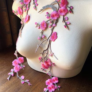 以下が含まれることがあります： 鮮やかなピンク色の刺繍入り花のアップリケ。詳細な花と繊細な枝が特徴です。マネキンに展示されており、衣服の装飾としての可能性を示しています。デザインにはグレーのアクセントと複雑なステッチが含まれています。