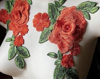 May include: Orange and green embroidered floral applique with three large flowers and several smaller flowers. The applique is on a white background.