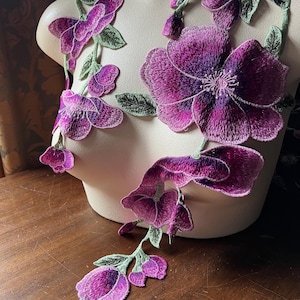 May include: Purple and green floral embroidery applique with a large flower and smaller flowers on a vine.