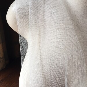 May include: Off-white tulle fabric draped over a mannequin bust. The tulle has a fine, textured weave, creating a delicate, semi-transparent appearance. The fabric is folded and layered, showcasing its soft, flowing quality. Ideal for bridal veils or other craft projects.