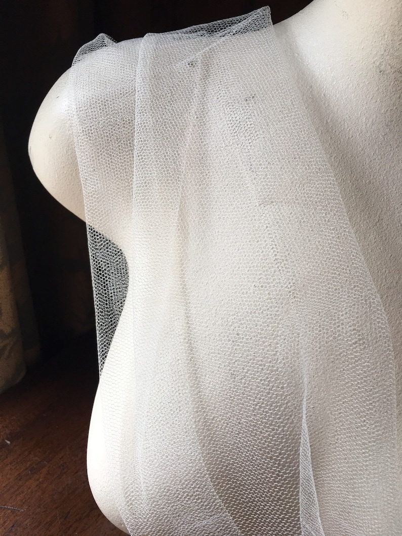May include: Off-white tulle fabric draped over a mannequin bust. The tulle has a fine, textured weave, creating a delicate, semi-transparent appearance. The fabric is folded and layered, showcasing its soft, flowing quality. Ideal for bridal veils or other craft projects.