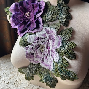 May include: Two embroidered fabric flowers, one purple and one white with purple accents, with green leaves, on a white mannequin.
