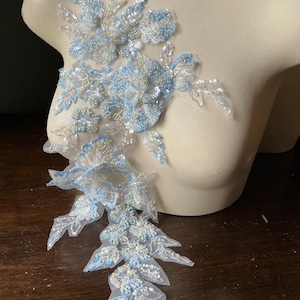 May include: A delicate, floral applique featuring light blue and silver sequins and beads. The design includes multiple flower and leaf shapes, with a sheer, white mesh backing. The applique is displayed on a mannequin.