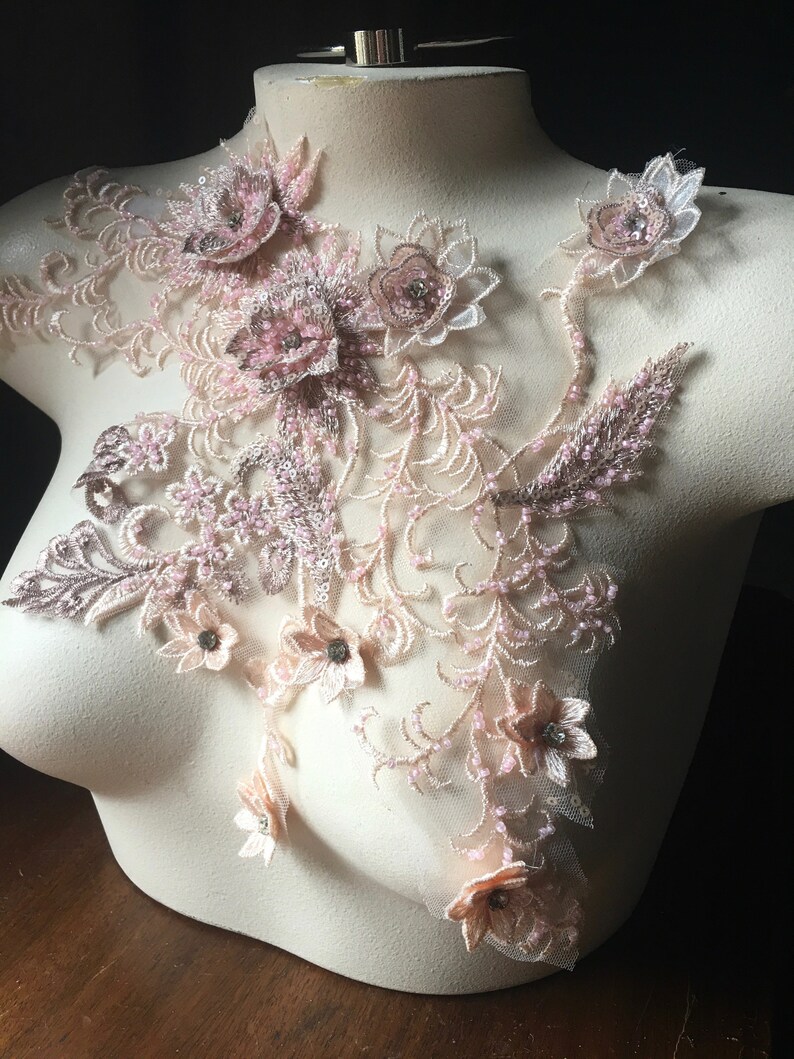 May include: A delicate floral lace applique with pink and beige flowers and beads. The applique features a variety of floral designs, including roses, lilies, and daisies. The applique is perfect for adding a touch of elegance to any garment or project.