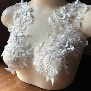 May include: Two white lace appliques with silver embroidery, featuring floral designs with leaves and delicate flowers. The appliques are perfect for adding a touch of elegance to wedding dresses, bridal accessories, or other craft projects.