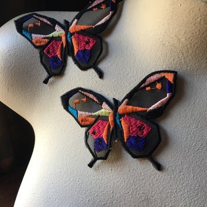 Puede incluir: Dos parches de mariposa de colores para planchar con contornos negros y un fondo de malla transparente. Las mariposas tienen un diseño geométrico con tonos de rosa, naranja, amarillo, azul y morado.