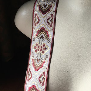 May include: Decorative ribbon with a repeating floral pattern in red, gold, and brown on a light gray background. The ribbon has a red border and is displayed against a mannequin.