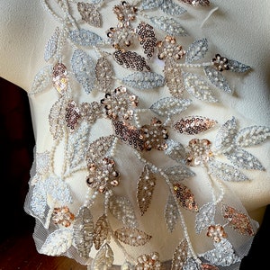 May include: A delicate floral applique featuring leaves and flowers in shades of silver, gold, and rose gold. The applique is made of sequins, beads, and embroidery on a sheer mesh fabric.