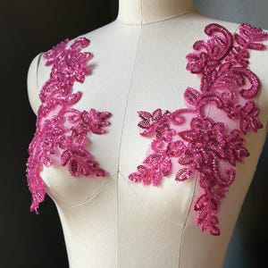 May include: Two magenta floral lace appliques with sequins. The appliques are designed with a leaf and swirl pattern. The appliques are displayed on a mannequin.