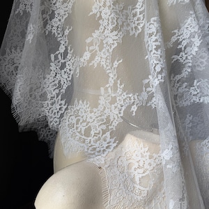 Könnte beinhalten: Ein weißer Spitzenstoff mit einem detaillierten Blumenmuster und gebogten Kanten. Das zarte, halbtransparente Material ist über eine Form drapiert und zeigt die filigrane Handarbeit sowie den feinen Netzgrund des Textildesigns.