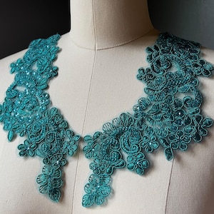 May include: Teal lace applique with floral and scroll designs. The applique is displayed on a mannequin, showcasing its intricate details and delicate craftsmanship. The lace is embellished with small, sparkling accents.