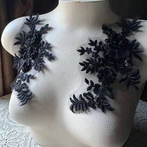 Può includere: Applicazioni in pizzo floreale nero esposte su un busto di manichino bianco. Le applicazioni presentano dettagliati disegni di foglie e fiori, attaccati a un supporto in rete trasparente. Il busto è posto su uno sfondo marrone scuro.