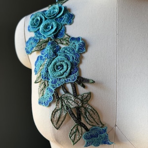 Puede incluir: Una aplicación floral bordada que presenta varias rosas en tonos azul cerceta, turquesa y azul real. El diseño incluye hojas y tallos detallados en tonos verdes y plateados, creando un arreglo vertical de flores de tela superpuestas.