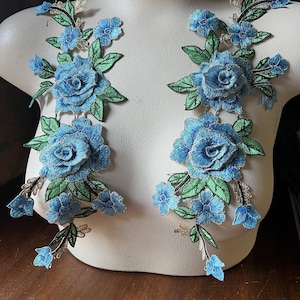 Puede incluir: Dos apliques florales azules y verdes con detalles plateados. Los apliques presentan rosas grandes y flores más pequeñas.