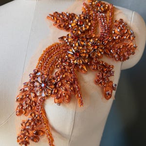 May include: An orange floral applique featuring intricate beadwork and faceted crystals on a sheer mesh backing. The design includes layered petals, sparkling amber-toned stones, and delicate embroidery arranged in a flowing, organic shape.
