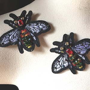 May include: Two embroidered bee patches. The bees have black bodies, detailed wings, and are embellished with red, gold, and green rhinestones. The patches are on a light-colored surface.