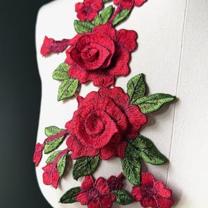 以下が含まれることがあります： 刺繍されたバラと緑の葉が特徴の鮮やかな赤い花のアップリケ。マネキンに表示されており、ファッションやクラフトプロジェクトでの使用が示唆されています。