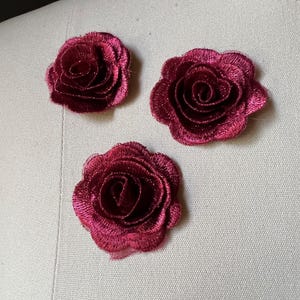 Puede incluir: Tres apliques de rosas burdeos bordadas. Las rosas están en plena floración, con costuras detalladas que crean una apariencia texturizada. Los apliques están dispuestos sobre una tela beige claro, mostrando su color vibrante y su intrincado diseño.