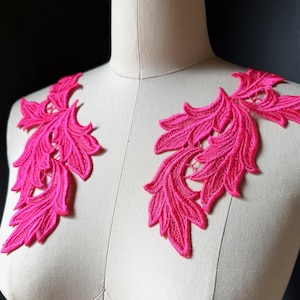 以下が含まれることがあります： 鮮やかなホットピンクの刺繍レースアップリケ2枚。葉と蔓の模様が詳細に施されています。これらの左右対称の装飾パーツは、ベージュの布製トルソーの胸元に配置されており、複雑なステッチと質感が際立っています。