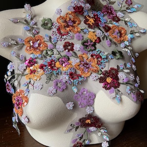 Puede incluir: Aplicación floral con una variedad de lentejuelas y cuentas de colores. El diseño presenta flores naranjas, rojas, moradas y azules con acentos verdes sobre un respaldo de malla transparente. La aplicación se muestra en un maniquí.