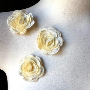 Puede incluir: Tres apliques de rosas de tela color crema. Cada flor presenta una construcción detallada de pétalos con un centro amarillo ligeramente más oscuro. Los apliques son probablemente para embellecer ropa o manualidades.