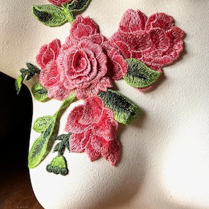 Pode incluir: Aplique de rosa vermelha bordada com folhas verdes. O desenho floral é fixado a uma superfície creme. O aplique apresenta costuras detalhadas e uma aparência realista, ideal para embelezar tecidos.
