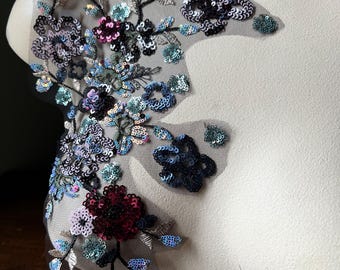 May include: A floral applique with a black mesh backing. The applique features a variety of flowers in shades of blue, purple, and burgundy, embellished with sequins. The design includes leaves and stems, creating a delicate, decorative piece.