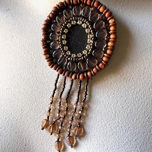 May include: A circular brooch featuring a central black disc surrounded by a ring of clear faceted beads, a border of small metallic beads, and an outer edge of brown wooden beads. Five beaded strands with clear brown beads hang below.