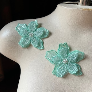 Może przedstawiać: Dwa jasnozielone aplikacje kwiatowe z koralików z perłowym środkiem. Aplikacje znajdują się na białym tle.