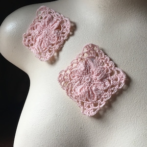 May include: Two light pink, square crochet appliques. Each features a floral design at the centre, surrounded by an openwork border. The appliques are displayed on a mannequin, showcasing their delicate texture and colour.