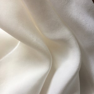 May include: Close-up of a flowing, off-white fabric with subtle texture. The material drapes in soft folds, creating shadows and highlights. The fabric appears to be lightweight and possibly silk or a similar material.