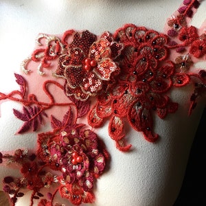 Puede incluir: Aplicación de encaje floral rojo con lentejuelas y cuentas. La aplicación presenta bordados detallados en varios tonos de rojo, incluyendo una flor central con lentejuelas y un grupo de cuentas. El diseño está sobre un fondo de malla transparente.