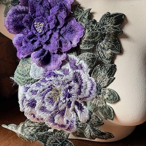 May include: Purple and white embroidered flower applique with green leaves. The applique is made of fabric and is perfect for adding a touch of elegance to any project.