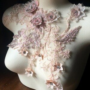 May include: A delicate floral lace applique with pink and beige flowers and leaves. The applique is adorned with small, clear rhinestones and beads.