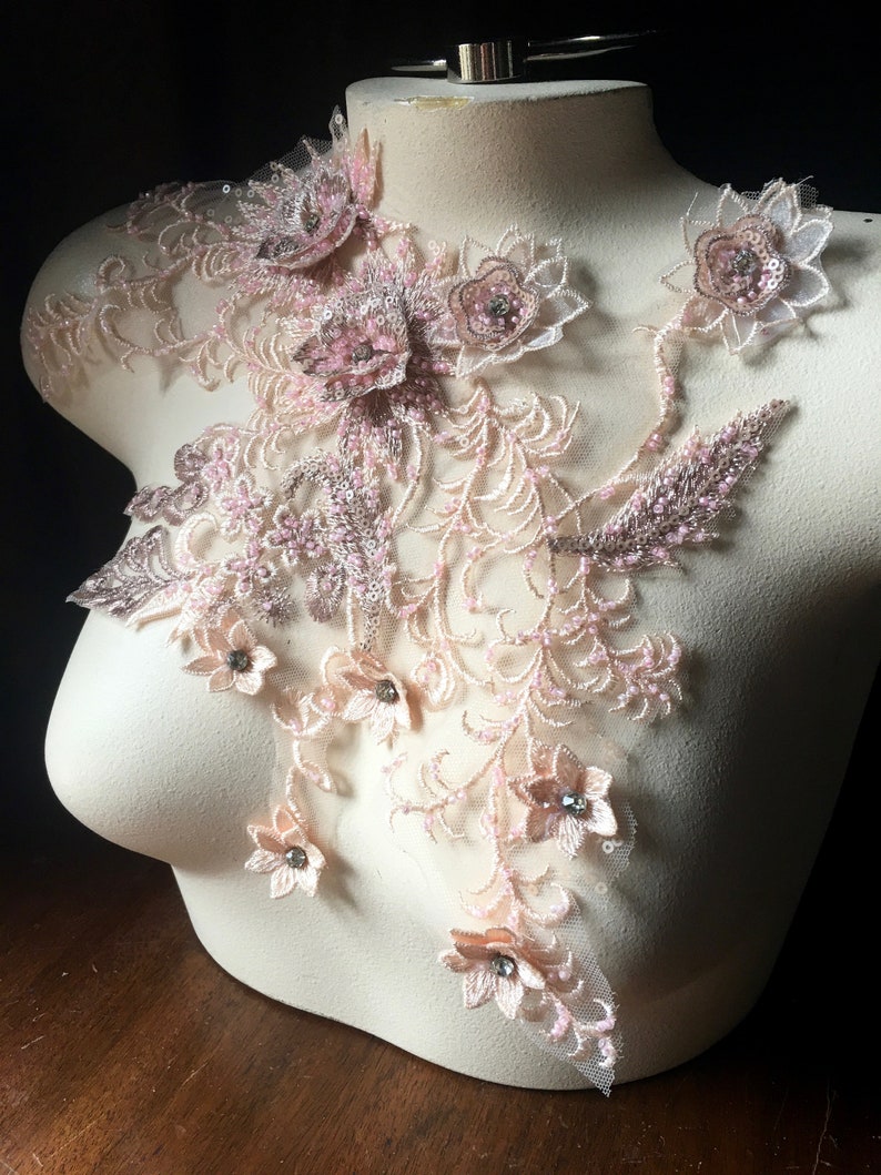 May include: A delicate floral lace applique with pink and beige flowers and leaves. The applique is adorned with small, clear rhinestones and beads.
