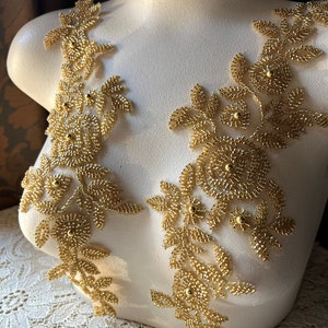 Könnte beinhalten: Zwei goldene Perlen-Blumenapplikationen auf einer weißen Schaufensterpuppe. Die Applikationen bestehen aus kleinen goldenen Perlen und haben ein zartes, filigranes Design.