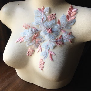 Może przedstawiać: Kwiatowa aplikacja z haftowanymi detalami w kolorze jasnoniebieskim i różowym. Aplikacja posiada centralny kwiat z różowymi koralikami oraz delikatne liście i gałązki. Aplikacja jest prezentowana na manekinie.