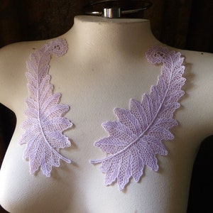 May include: Two delicate lilac lace appliques in the shape of a fern leaf. The appliques are perfect for adding a touch of elegance to any project.