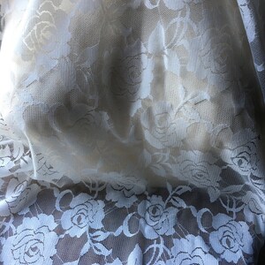 May include: Close-up of white lace fabric with a floral rose pattern. The delicate lace is semi-sheer, allowing light to filter through. The roses are intricately detailed, creating a romantic and elegant texture. Ideal for bridal wear, lingerie, or decorative projects.