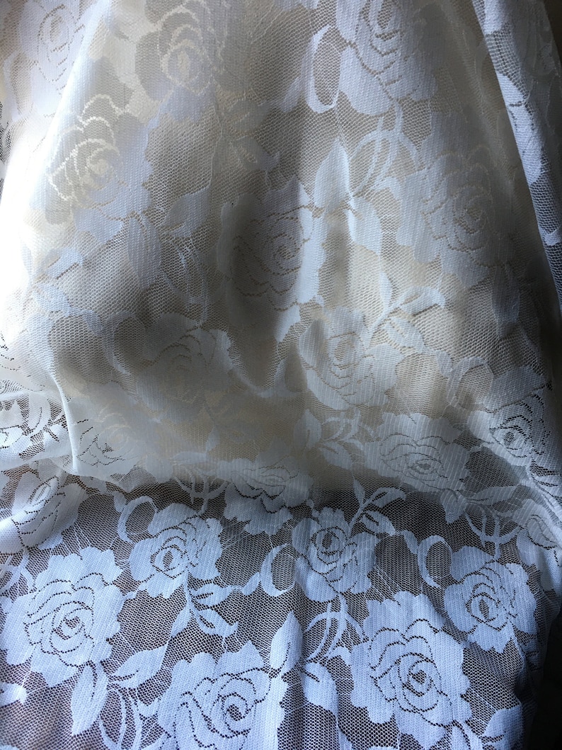May include: Close-up of white lace fabric with a floral rose pattern. The delicate lace is semi-sheer, allowing light to filter through. The roses are intricately detailed, creating a romantic and elegant texture. Ideal for bridal wear, lingerie, or decorative projects.
