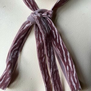 Puede incluir: Una corbata de terciopelo malva con un patr&oacute;n texturizado. La corbata est&aacute; anudada en el cuello y tiene extremos largos y fluidos. La tela tiene un aspecto suave y brillante, y el color es un p&uacute;rpura apagado.