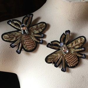 May include: Two embroidered bee appliques. Each bee features a black velvet base, gold metallic thread detailing, and a large, clear crystal at the center. The wings are intricately designed, and the body is filled with small, gold beads.