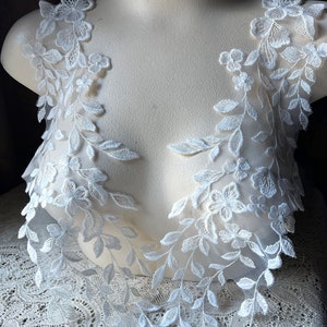 Può includere: Due applicazioni in pizzo bianco con motivi floreali e foglie. Le applicazioni sono su un busto di manichino bianco.