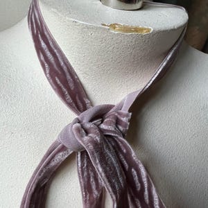 Puede incluir: Una corbata de terciopelo rosa empolvado est&aacute; anudada en el centro, con extremos largos y fluidos. La corbata est&aacute; colocada sobre un busto de maniqu&iacute; con una percha de metal plateado en la parte superior. El terciopelo tiene un brillo sutil.