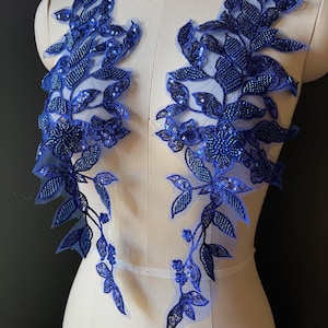 Könnte beinhalten: Zwei marineblaue Blumenapplikationen mit Perlen- und Pailletten-Details. Die Applikationen weisen ein Blatt- und Blumendesign auf, das auf einem transparenten Träger befestigt ist. Die Applikationen werden auf einer Schaufensterpuppe präsentiert.