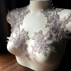 May include: Two delicate lace appliques in a light blush colour, featuring floral designs with intricate details. The appliques are displayed on a white dress form.