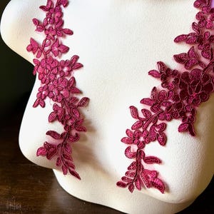 May include: Two burgundy floral lace appliques are displayed on a white mannequin bust. The appliques feature detailed floral and leaf patterns, ideal for embellishing garments or crafts. The lace is a deep, rich burgundy colour.