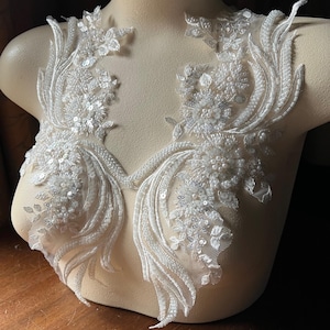 May include: Two white beaded lace appliques with pearl and sequin embellishments. The appliques are displayed on a white dress form.