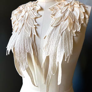May include: Two cream-coloured lace shoulder pieces, shaped like wings, are displayed on a mannequin. The lace has a detailed, feathery design, with layered textures and elongated, pointed tips. The background is a dark grey.