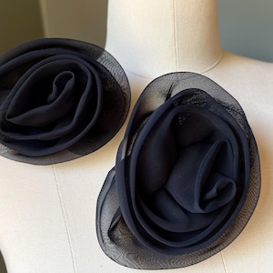 May include: Two navy blue fabric flower embellishments. Each flower is a layered design with a sheer, netted outer layer. The flowers are displayed on a mannequin, showcasing their shape and texture. These could be used as accessories.